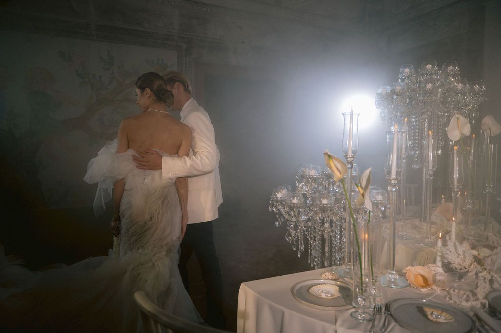 Bride and groom share romantic embrace near elegant crystal candelabra at dimly lit, atmospheric wedding reception.