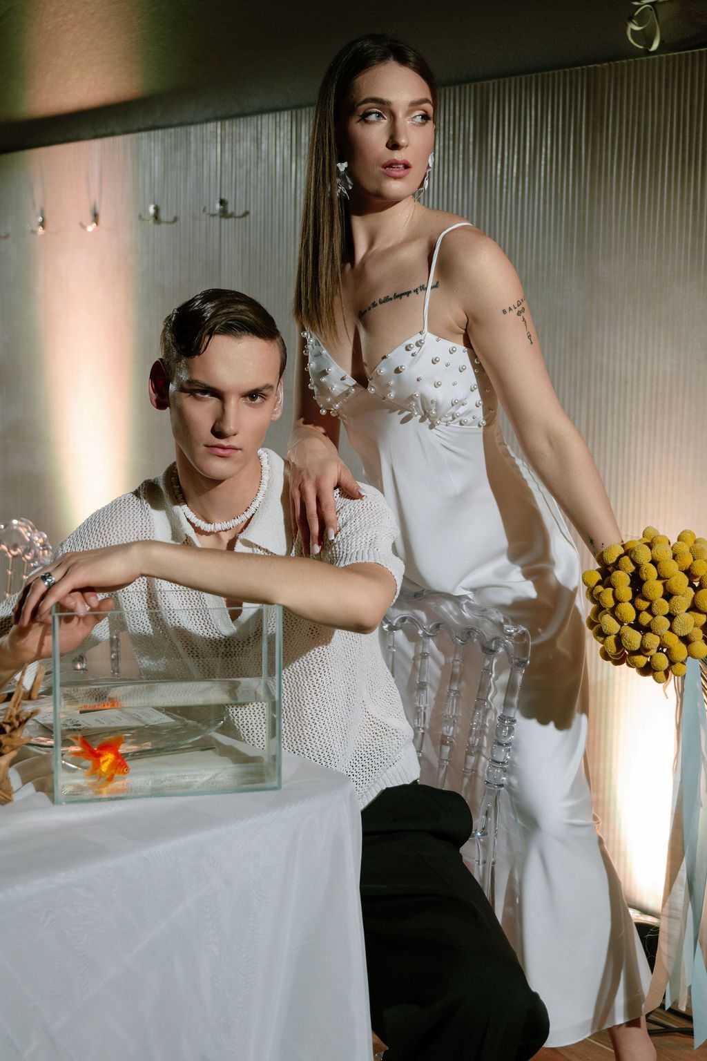 Two models pose elegantly, one in a white embellished dress beside a fish tank and yellow flowers.