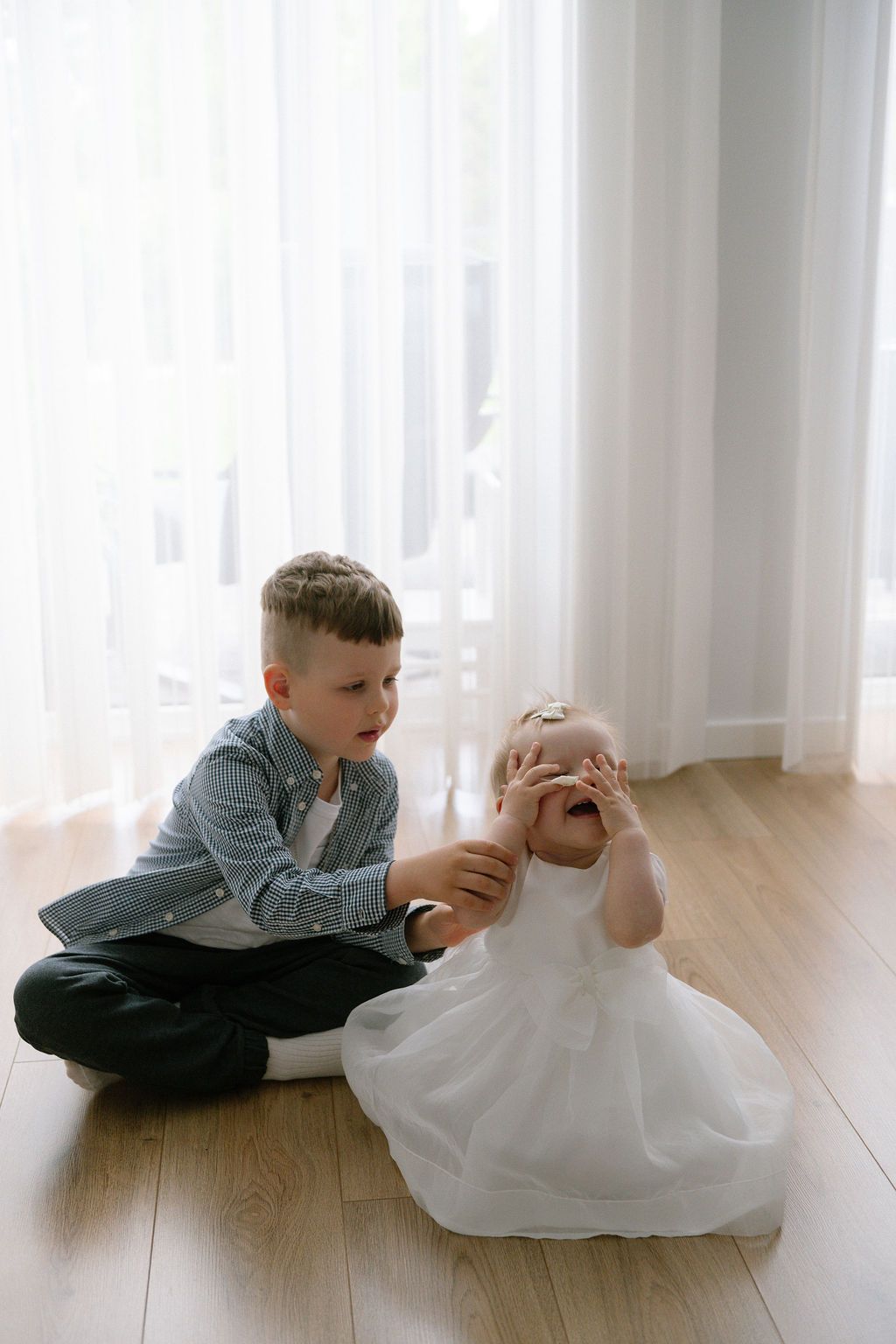 A young boy in a blue shirt sits on hardwood floor comforting a crying toddler girl in a white dress.