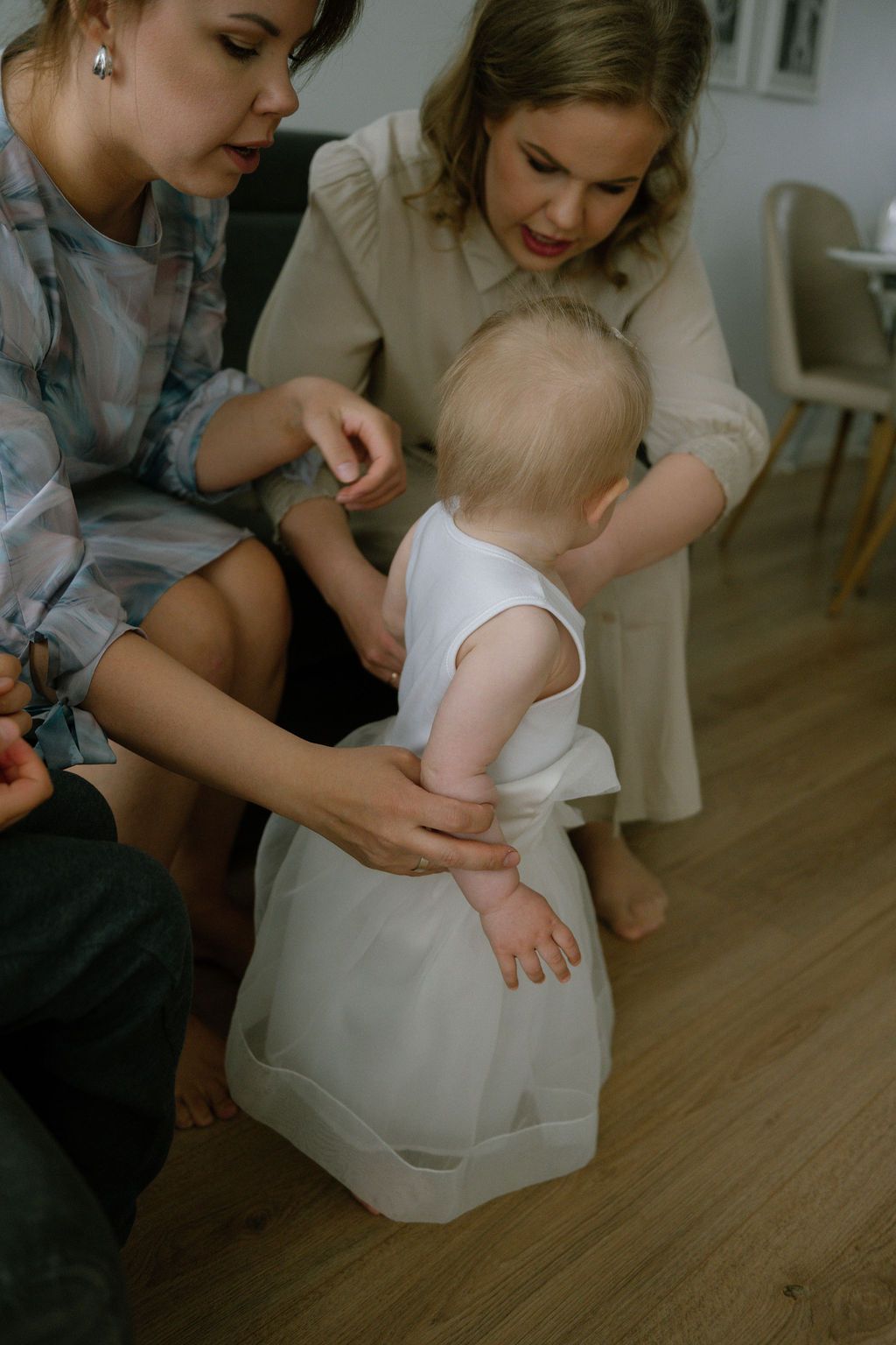 Two women help a baby in a white dress take her first steps on a wooden floor indoors.