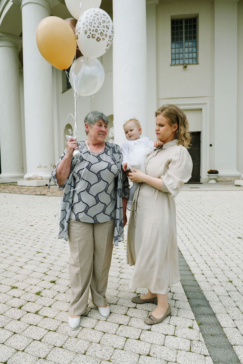 Grandmother and mother holding baby with gold and white balloons outside a white columned building.