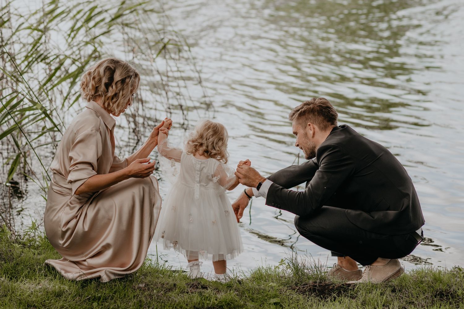 Family with toddler girl in white dress playing by a serene lakeside.