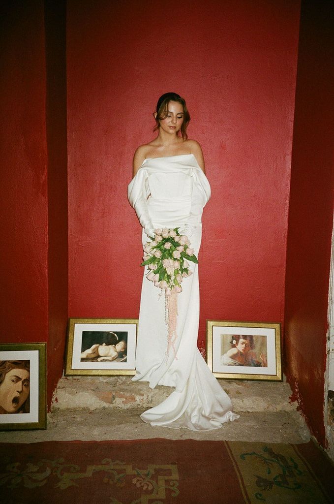 Bride in white off-shoulder gown holding pink bouquet, standing against red wall with framed artwork.