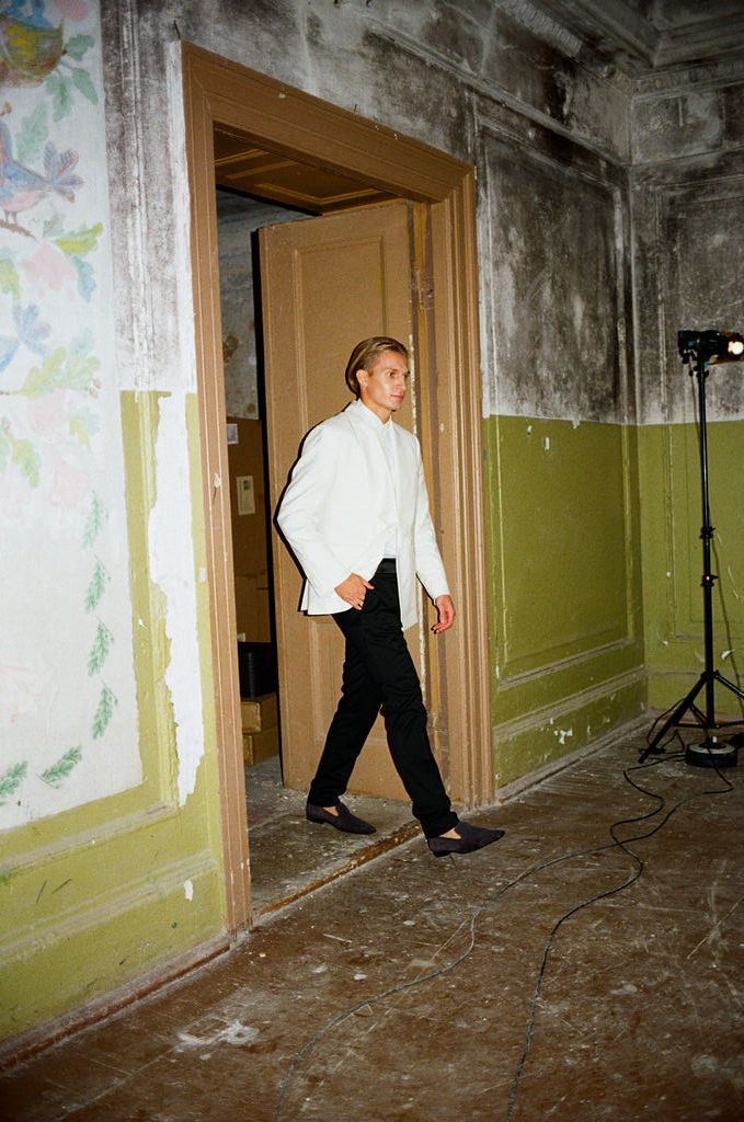 Young man in white shirt and black pants walking through doorway in rustic room with green walls.