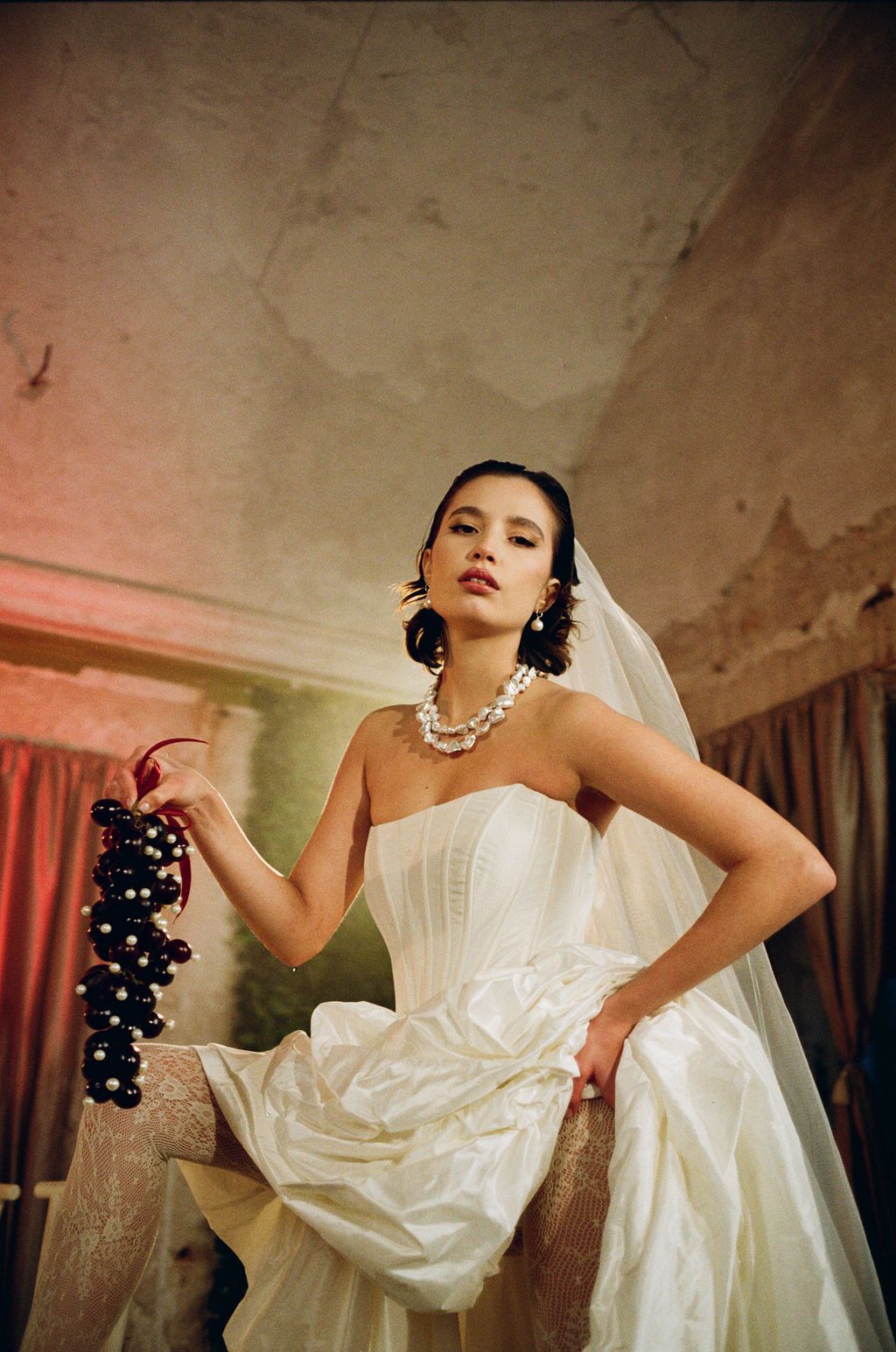 Bride in strapless white corset gown holding dark grapes, wearing pearl necklace and veil indoors.