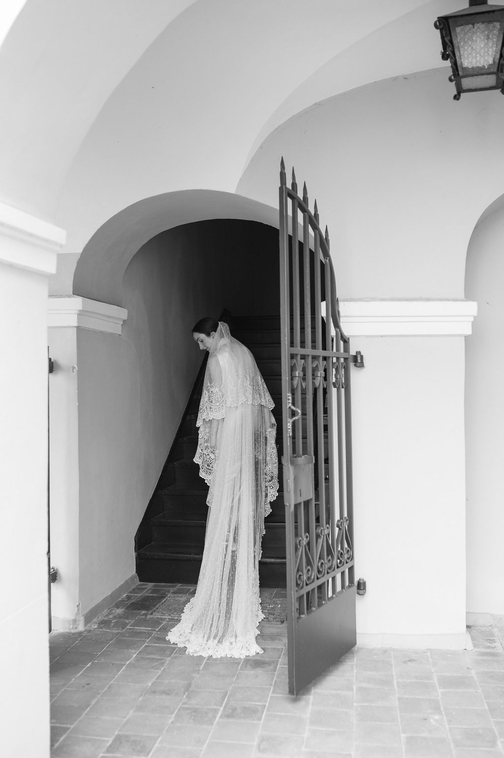 Bride in lace wedding gown and cathedral veil standing at ornate iron gate in black and white architectural setting.