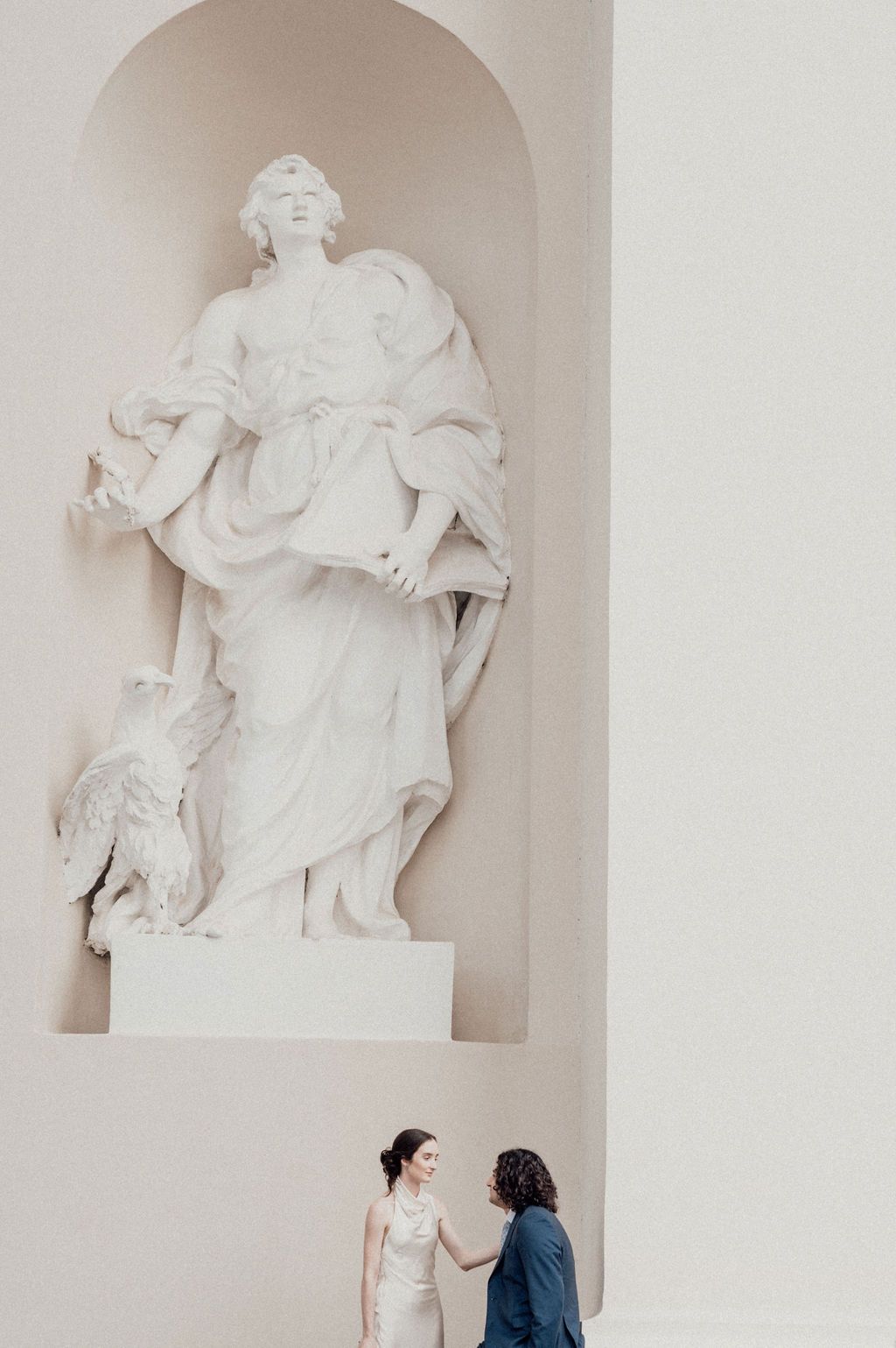 Bride and groom share a tender moment beneath an elegant white classical statue in a grand architectural niche.