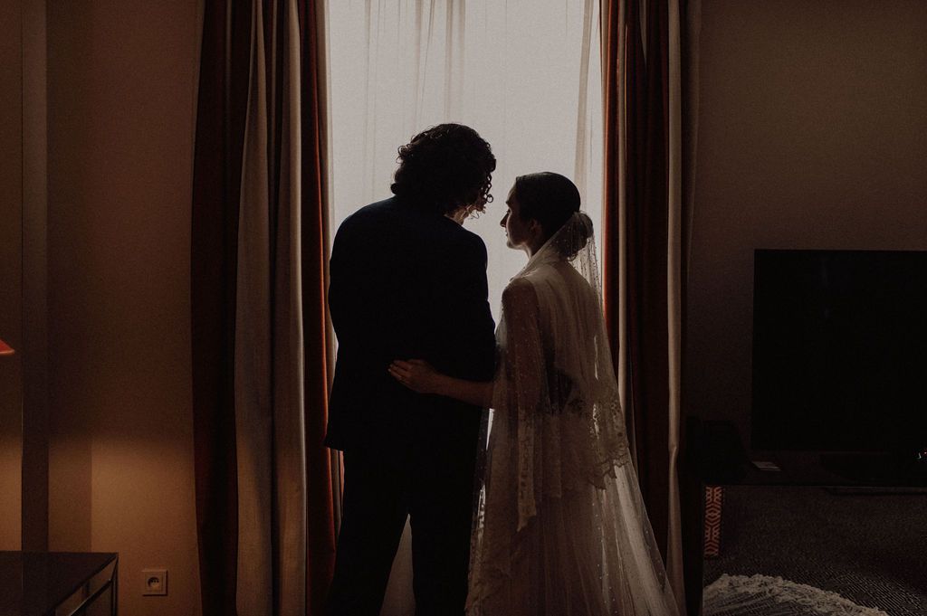 Silhouette of bride and groom standing together by a curtained window in a romantic indoor wedding setting.