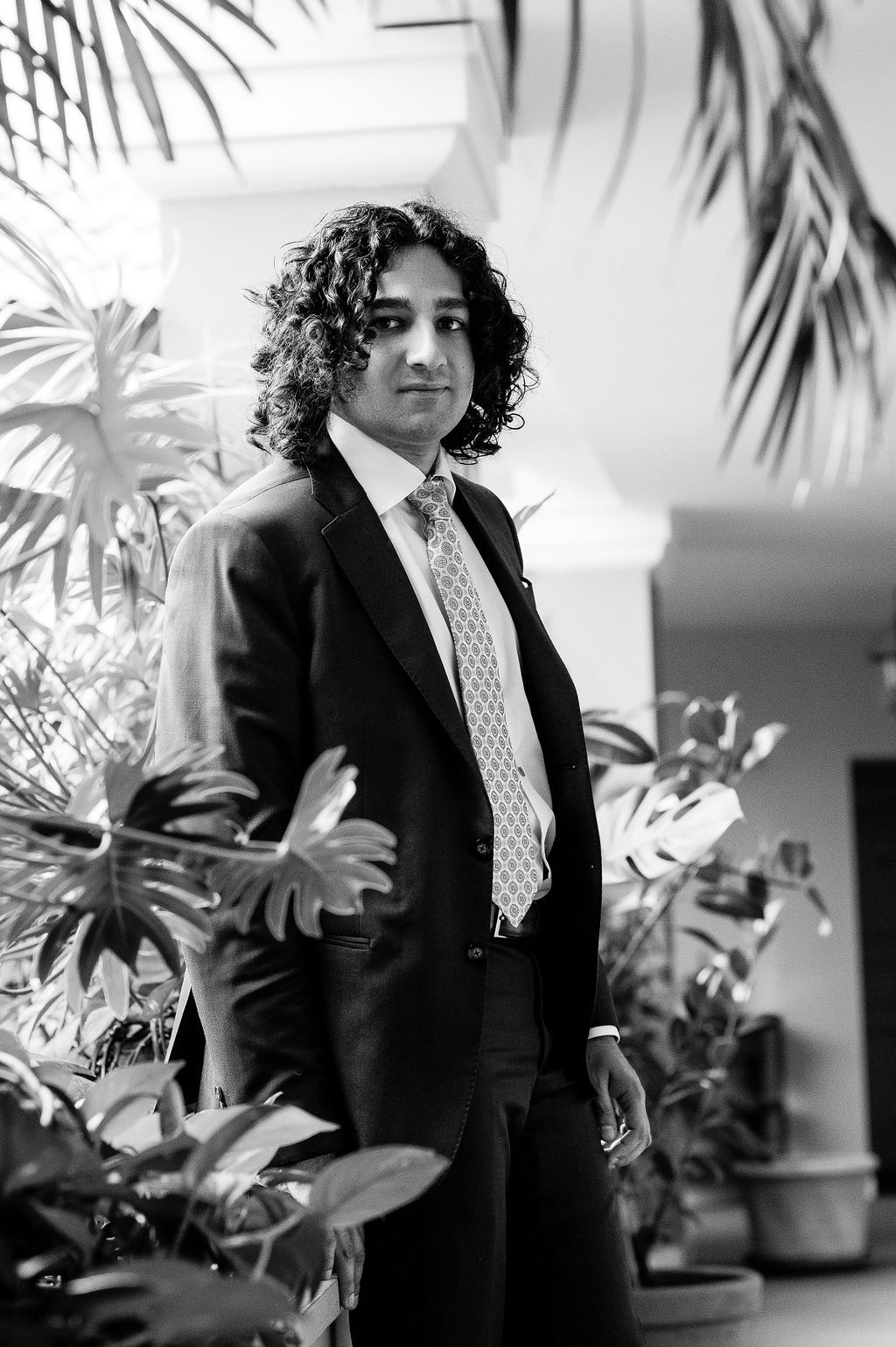 Young man with curly hair wearing a suit and tie poses among tropical plants in a black and white photo.