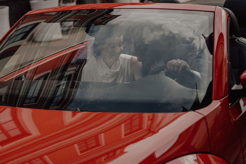 Two people conversing inside a red sports car, viewed through the windshield on a city street.