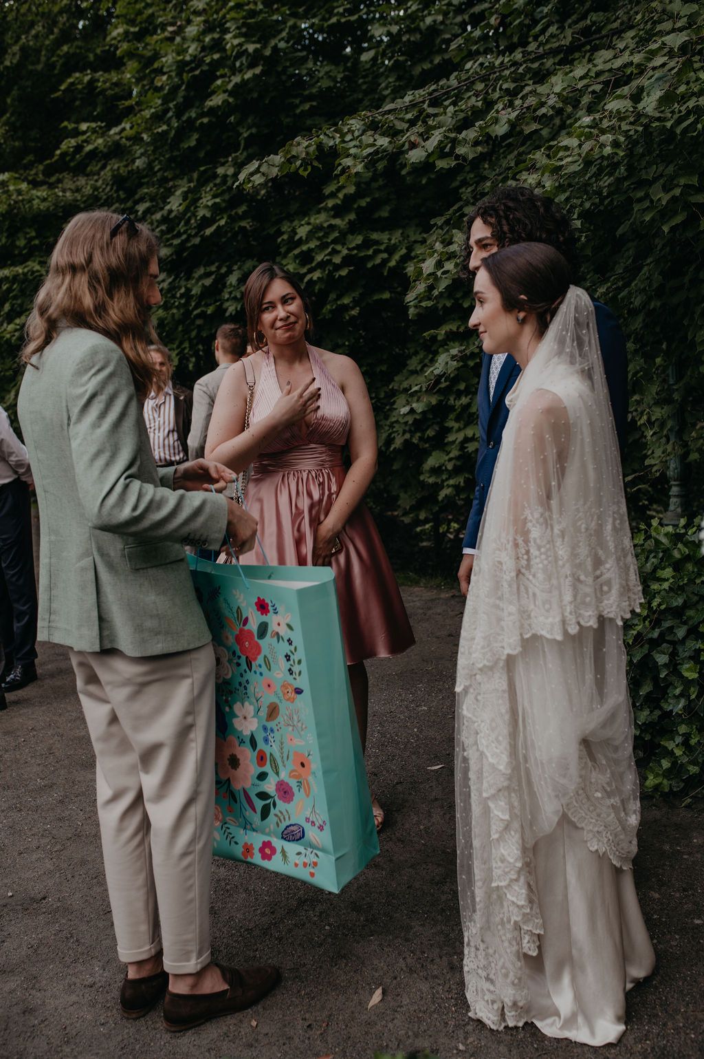 Wedding guests presenting a floral gift bag to a bride in lace veil and groom in navy suit outdoors.