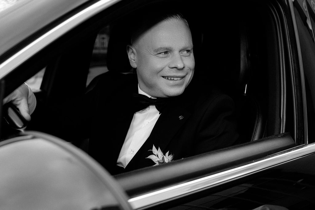 Smiling groom in black tuxedo with boutonniere sits in a luxury car, black and white wedding photo.