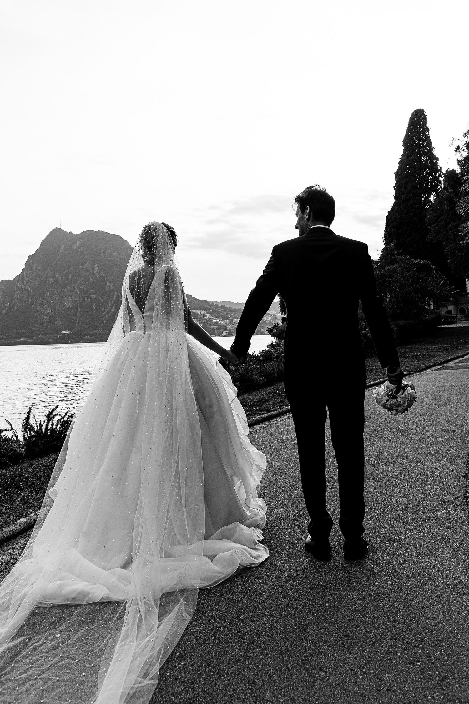 A bride and groom walk along a scenic mountain path in black and white, her flowing veil trailing behind them.