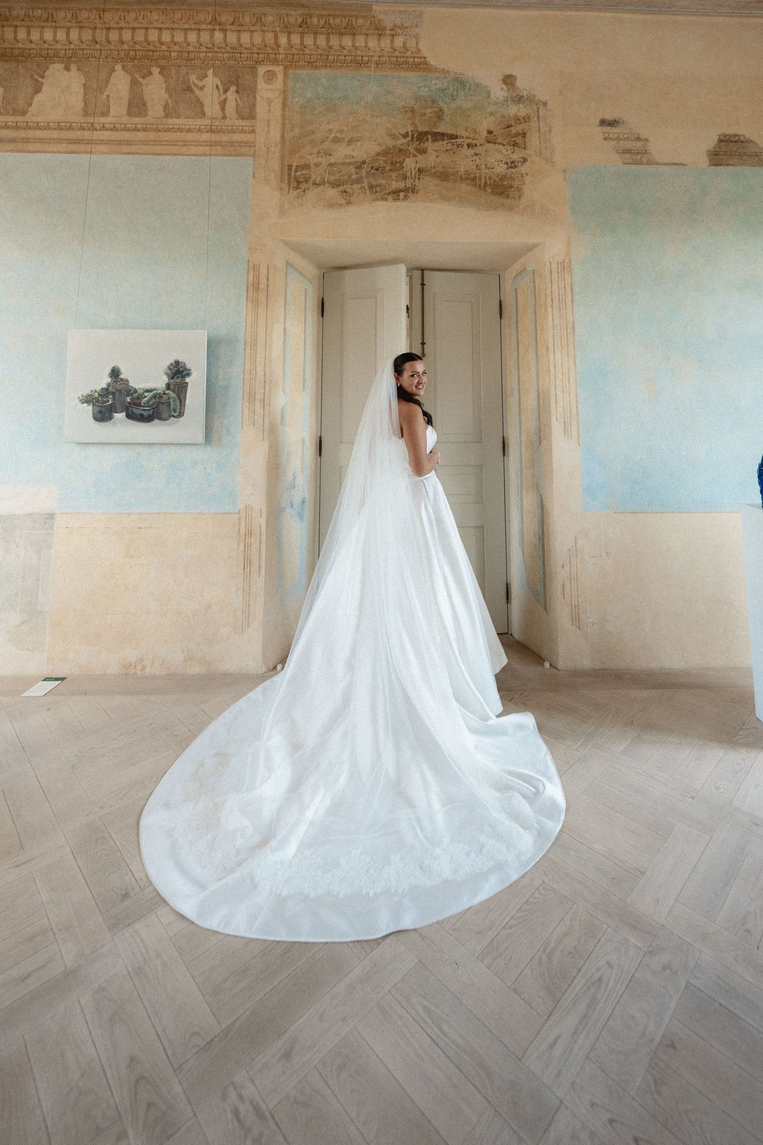 Bride in a white gown with a long cathedral train and veil stands in a historic room with frescoed walls and a herringbone wood floor.