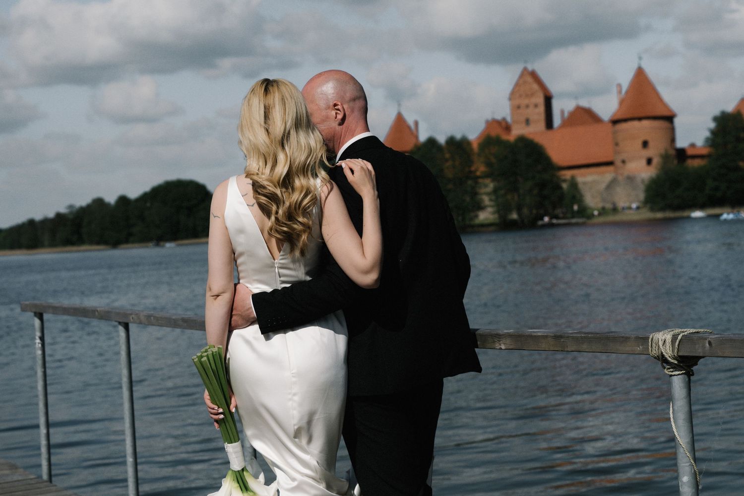 Bride in a white gown holding a bouquet embraces her groom on a waterfront pier with a red-roofed medieval castle across the lake.