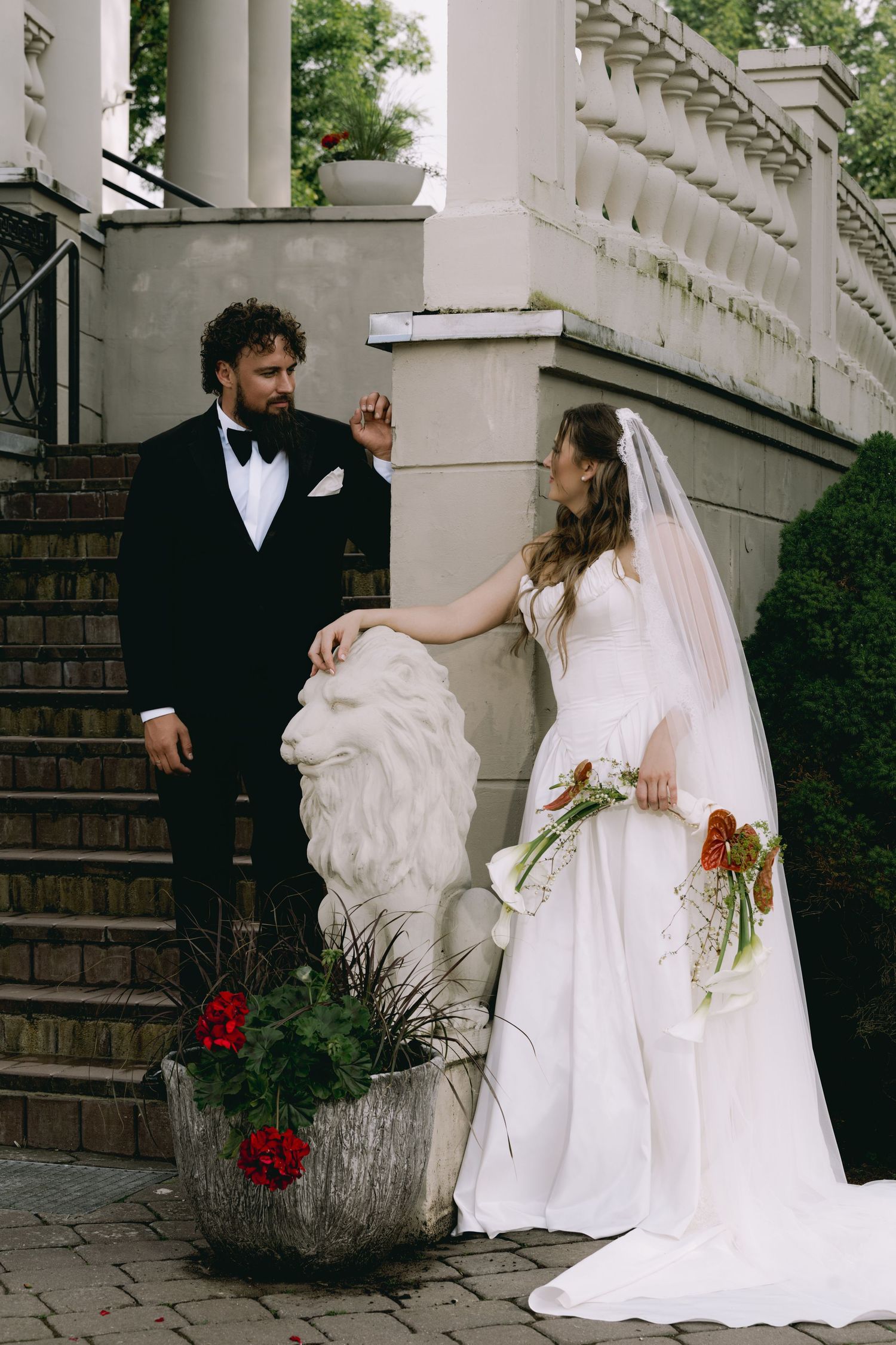 Bride and groom posing by stone lion sculpture on elegant staircase with red flowers.
