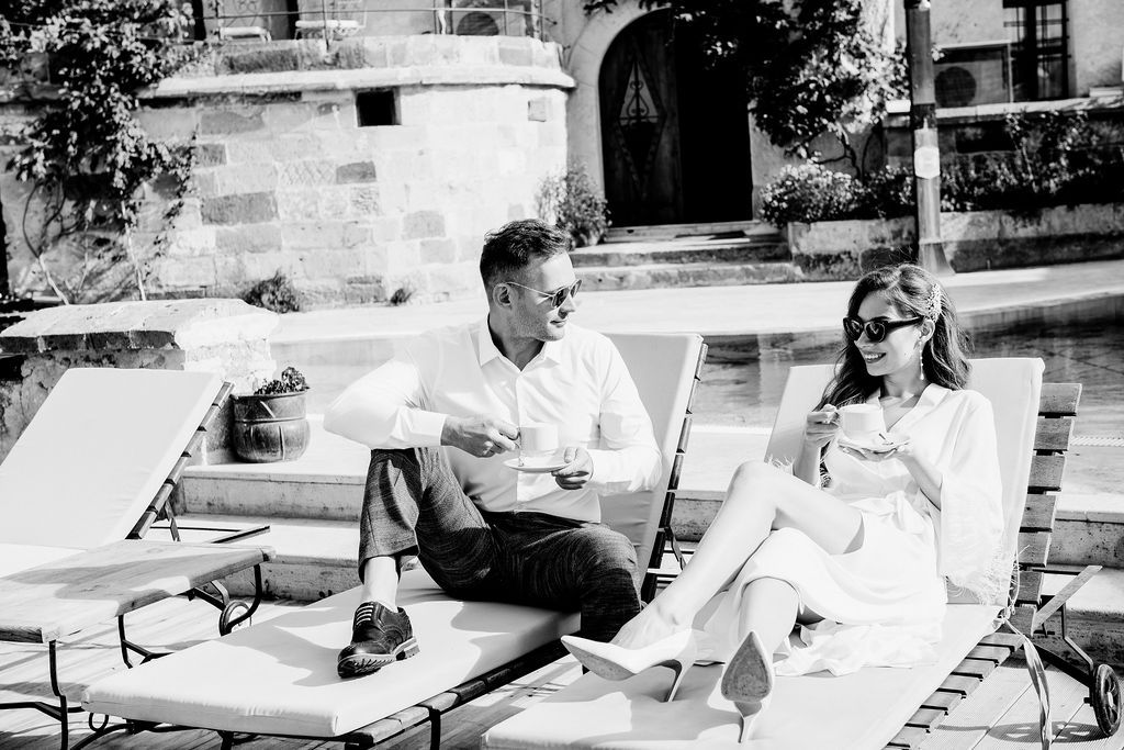 Couple relaxing on lounge chairs by a pool, enjoying coffee in a sunny outdoor setting.