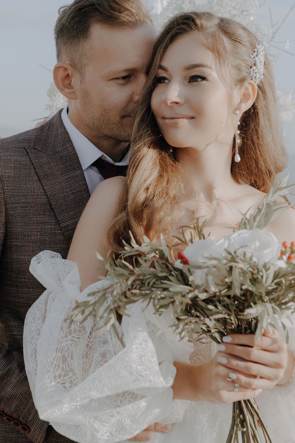 Groom kisses bride's cheek as she holds a lush bouquet with olive branches and white flowers.