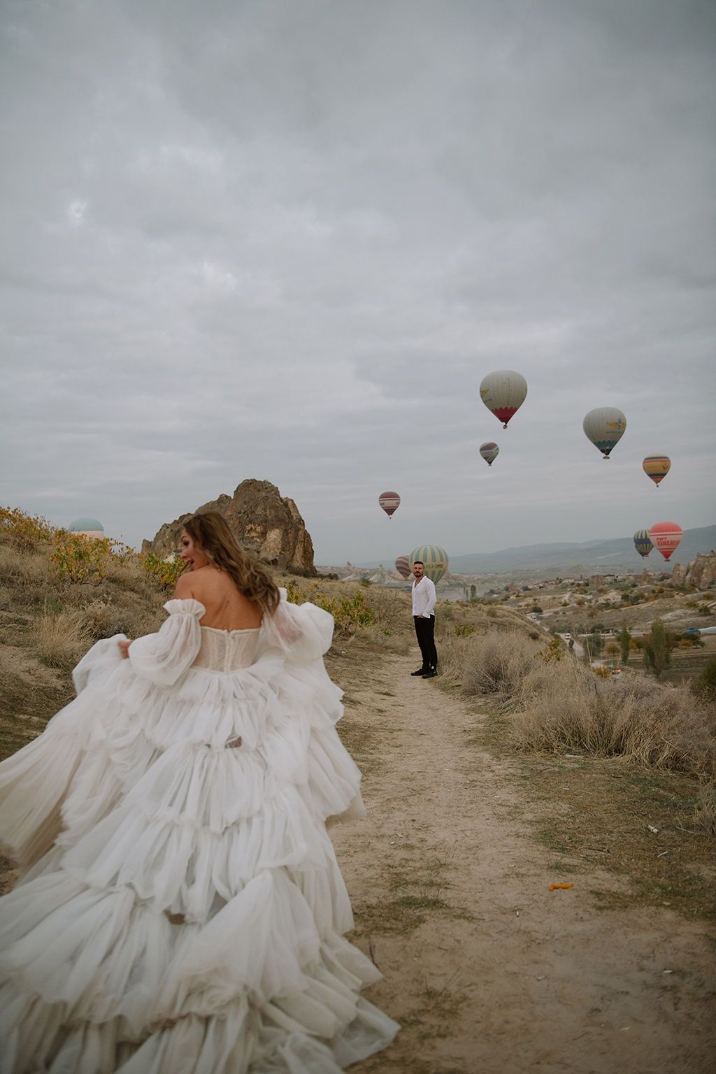 Bride in a ruffled white gown walks toward groom on a scenic Cappadocia path with hot air balloons in the cloudy sky.