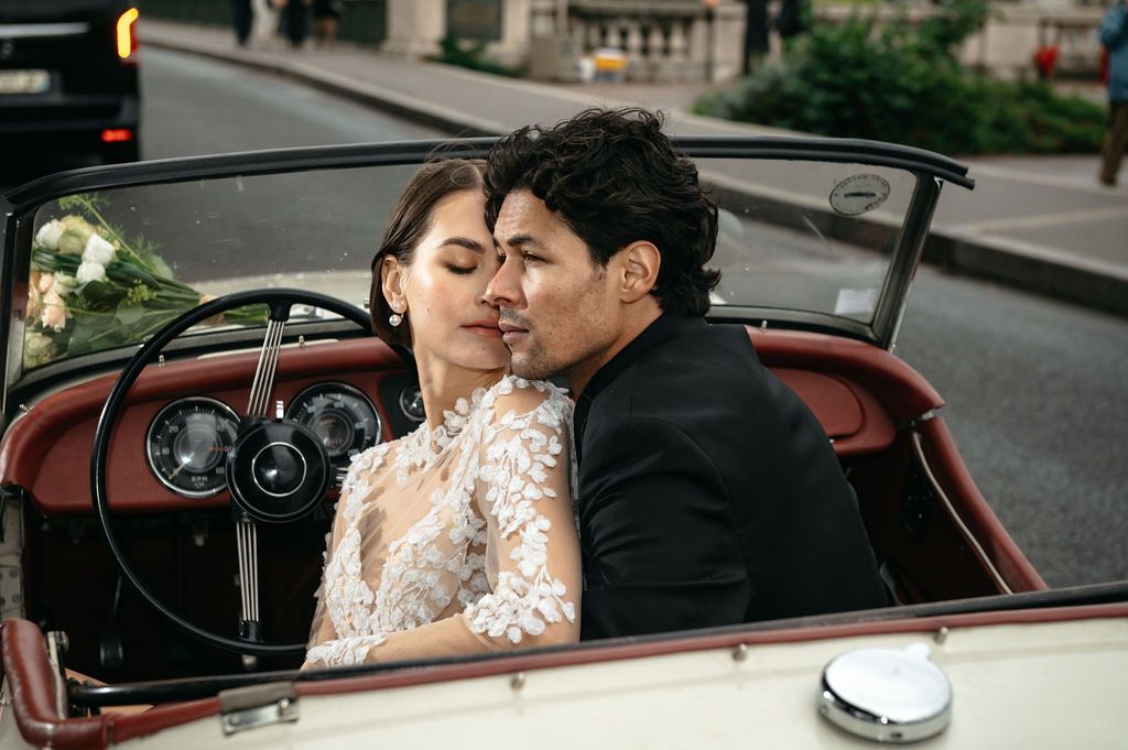 Couple sharing a romantic kiss in a vintage convertible car, bride in lace gown, groom in black suit.