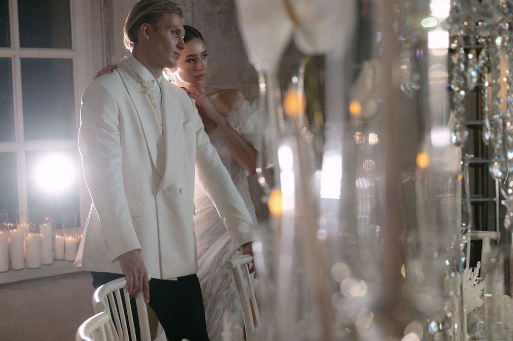 Elegant couple in white formal attire standing near crystal chandeliers in a softly lit, luxurious venue.
