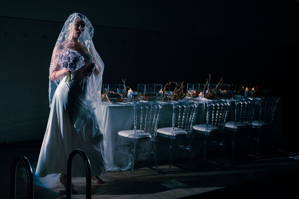 A bride in a white gown and veil stands beside a candlelit reception table with ghost chairs in dramatic dark lighting.