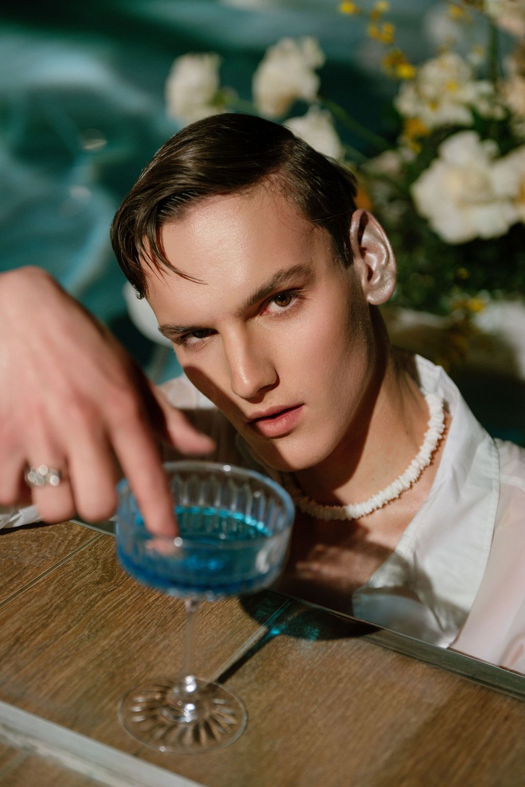Young man in white shirt and pearl necklace reaching for blue cocktail glass near white flowers.