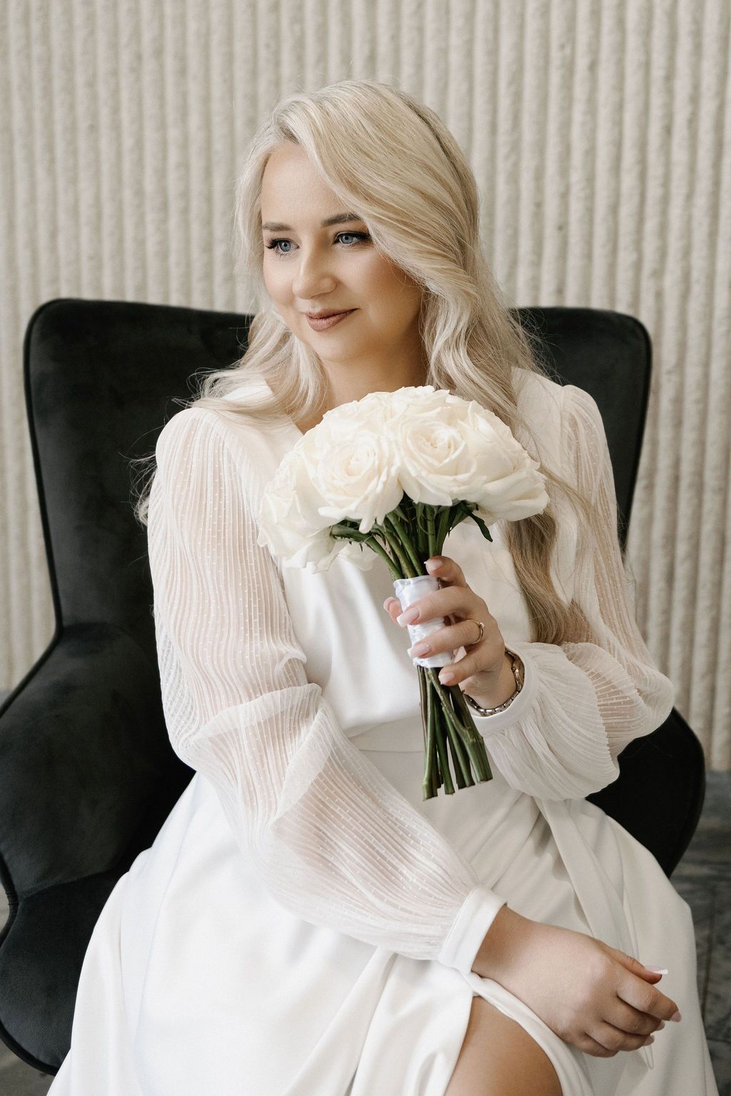 Blonde bride in white dress holding bouquet of white roses while seated in dark velvet chair.
