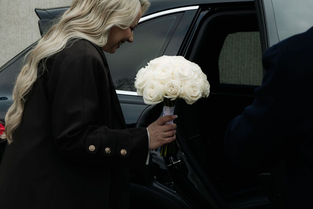 Blonde woman in black coat holding white rose bouquet while getting into a dark luxury car.