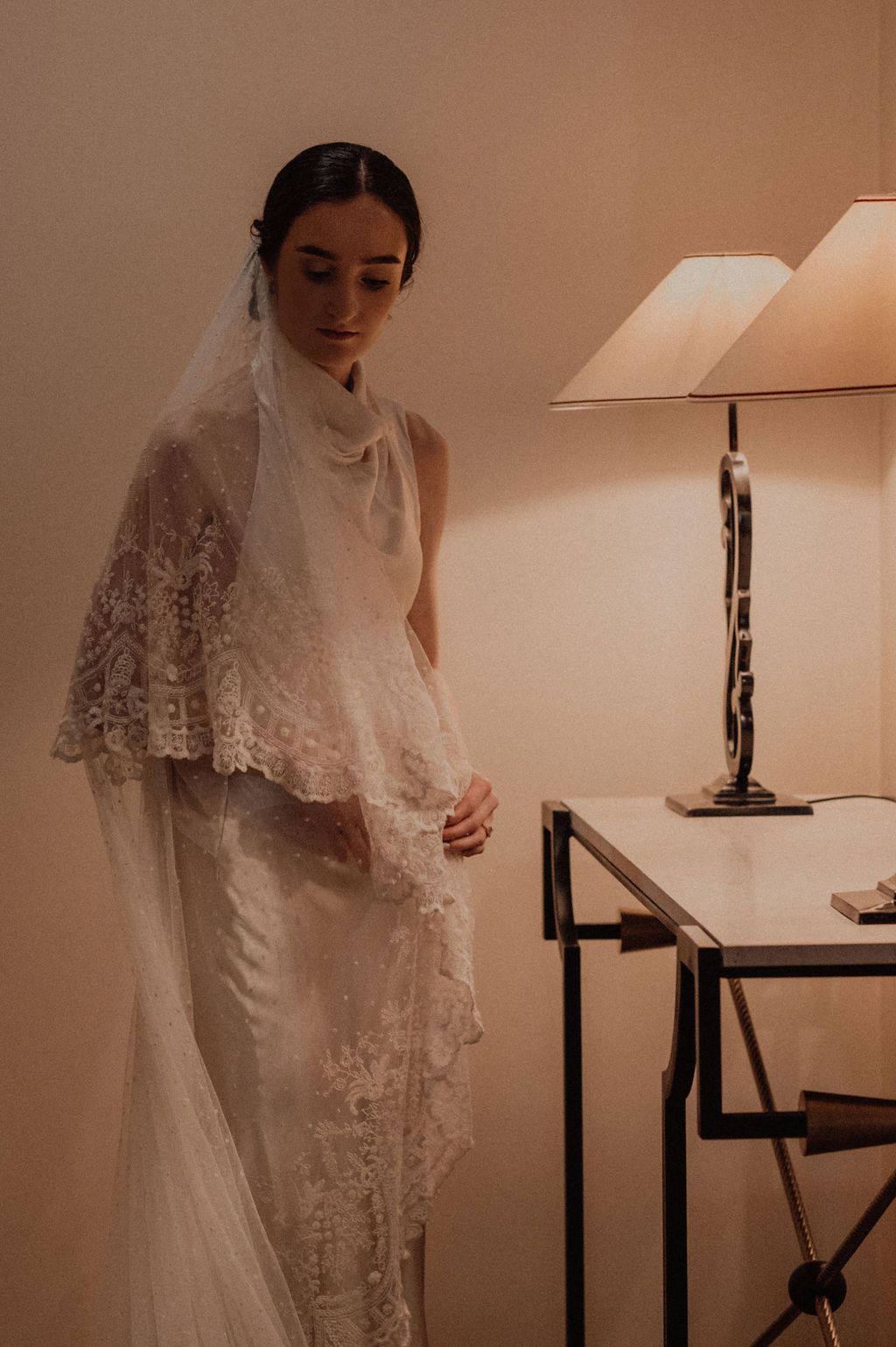 Bride in elegant lace wedding veil and gown standing by a lamp-lit table in a warmly lit room.