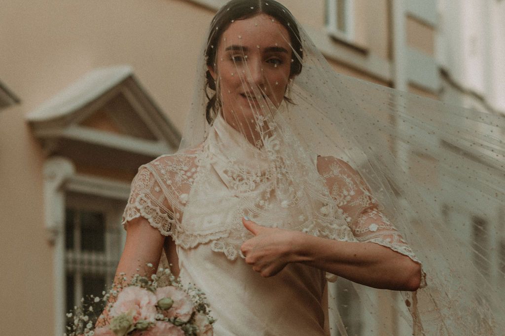Bride in lace wedding dress with flowing veil blowing in wind, holding pink bouquet outdoors.