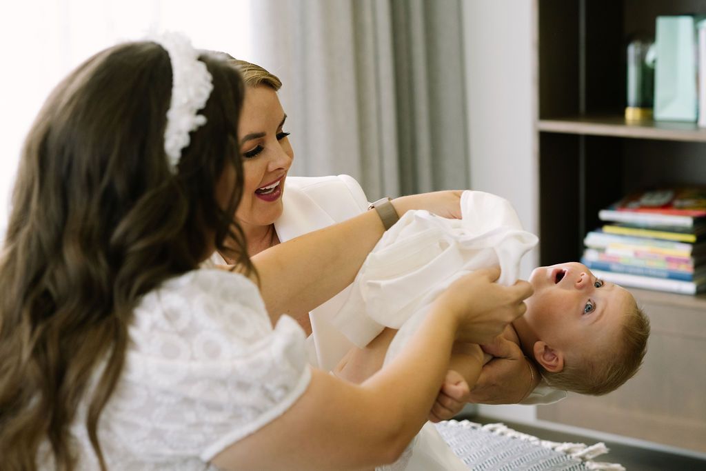 A smiling bride in a white dress holds a happy baby during a joyful wedding day getting-ready moment.