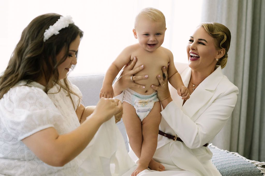 Two women in white outfits laugh joyfully while playing with a happy baby in a diaper indoors.
