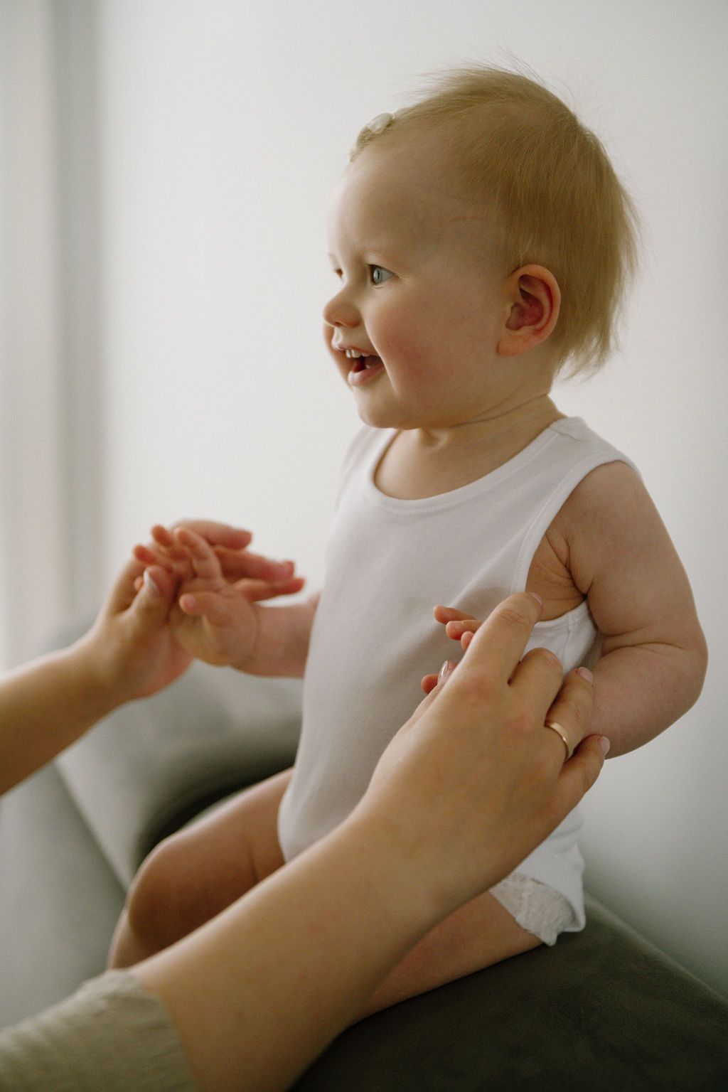 A happy smiling baby in a white onesie being held by a mother on a gray sofa.