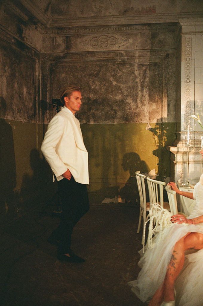 Groom in white jacket stands pensively beside a bride in a white dress in a rustic ornate venue.