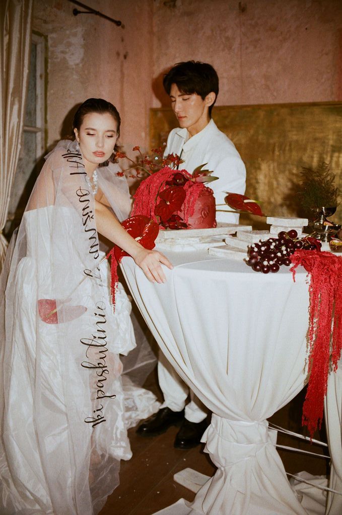 Couple in elegant wedding attire cutting a red floral cake at a draped table in a romantic vintage setting.