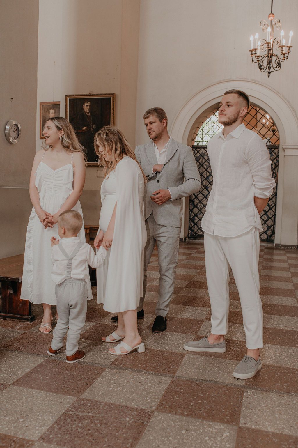Family dressed in white attire gathered inside an elegant church hall with chandelier and portrait painting.