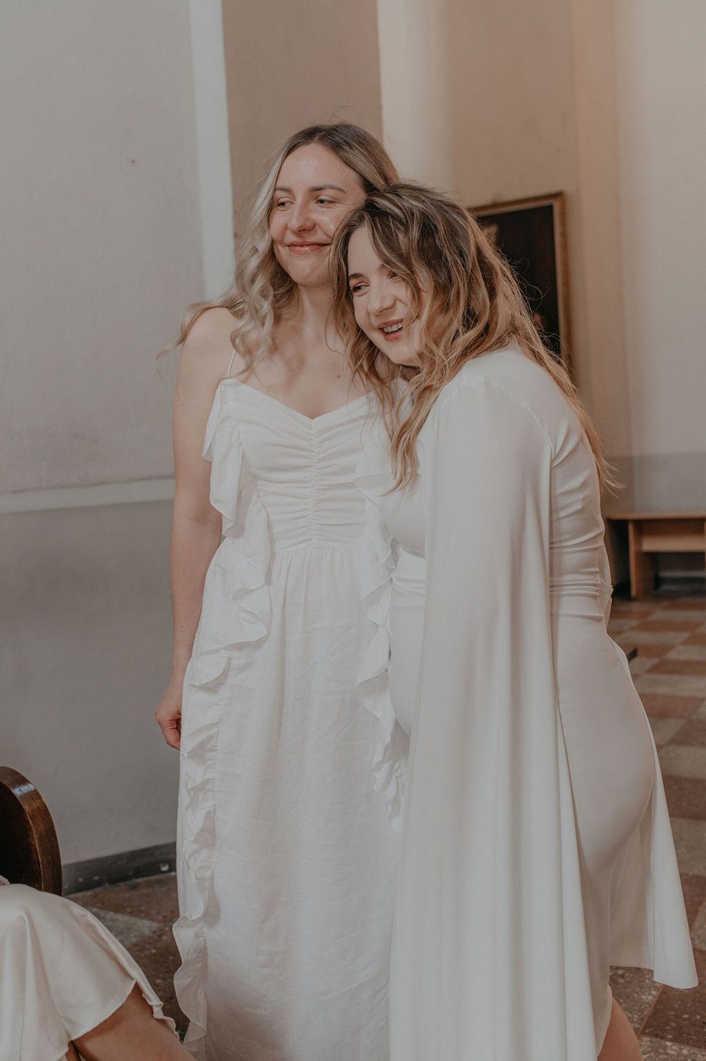 Two women in white dresses share a joyful embrace, smiling together in an elegant indoor setting.
