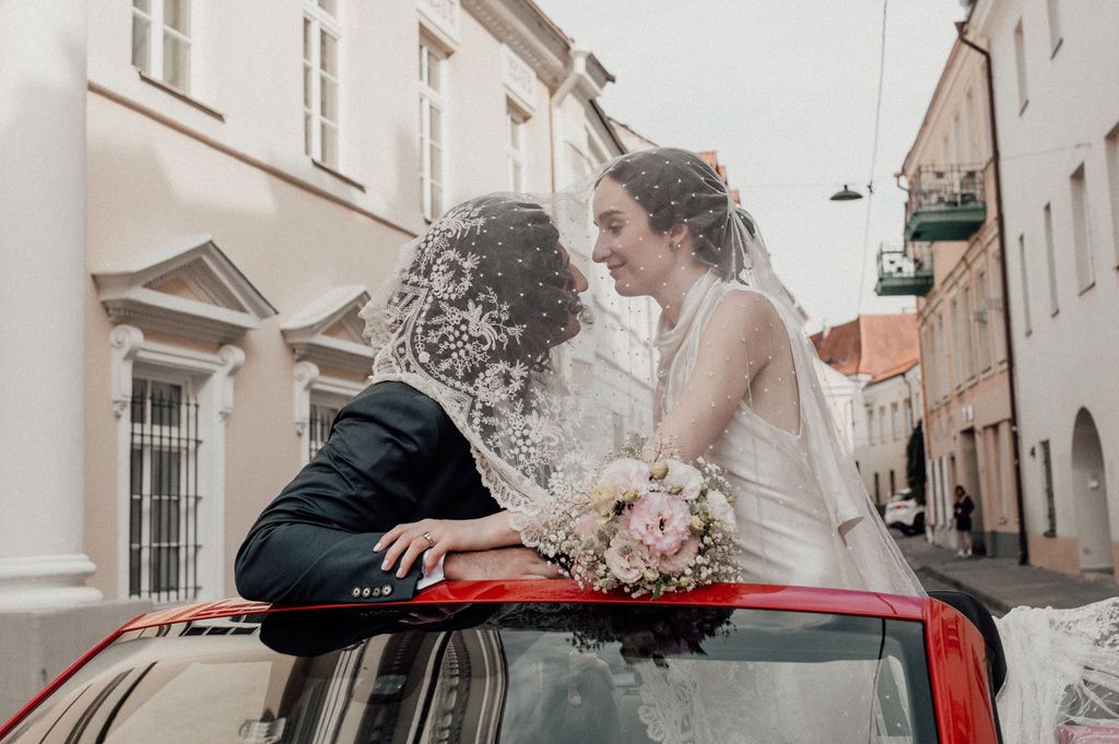 Bride and groom sharing a romantic kiss leaning out of a red car sunroof on a cobblestone street.