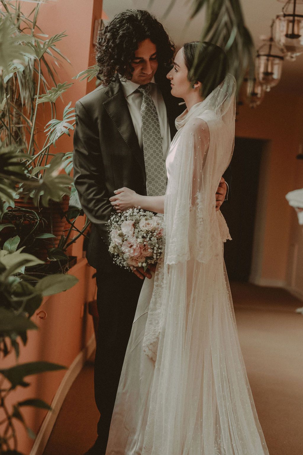 Bride and groom share an intimate moment indoors, she holds a bouquet in a lace gown with veil, he wears a dark suit.