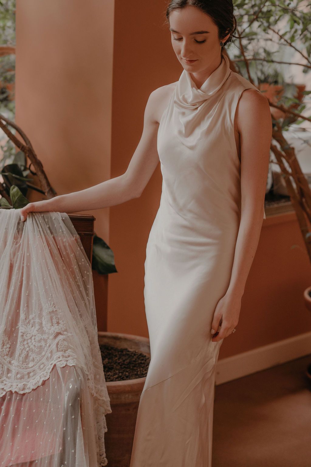 Bride in sleeveless ivory cowl-neck wedding gown holding a lace veil in a warm terracotta room.
