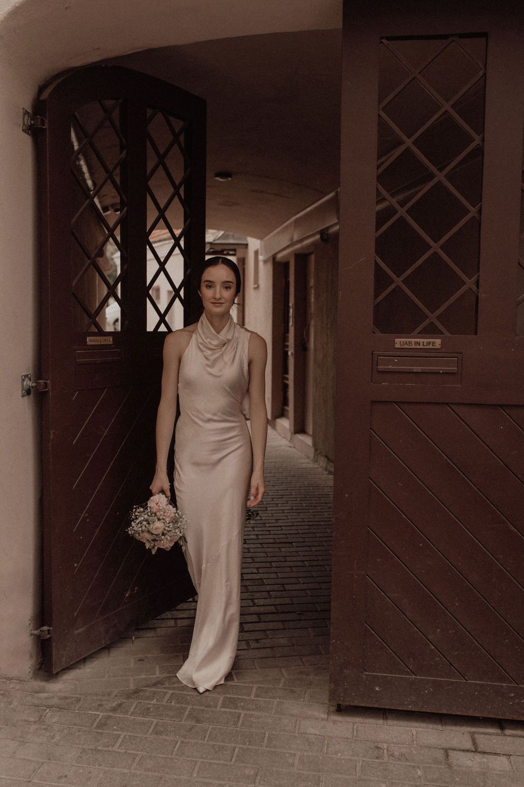 Bride in elegant champagne satin halter gown holding bouquet standing in doorway of rustic wooden doors.
