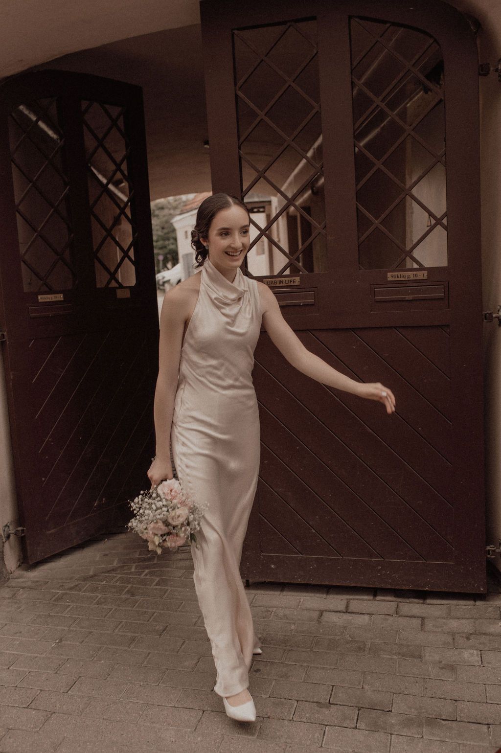 Bride in elegant ivory satin halter gown holding a pink bouquet, walking through ornate wooden doors.