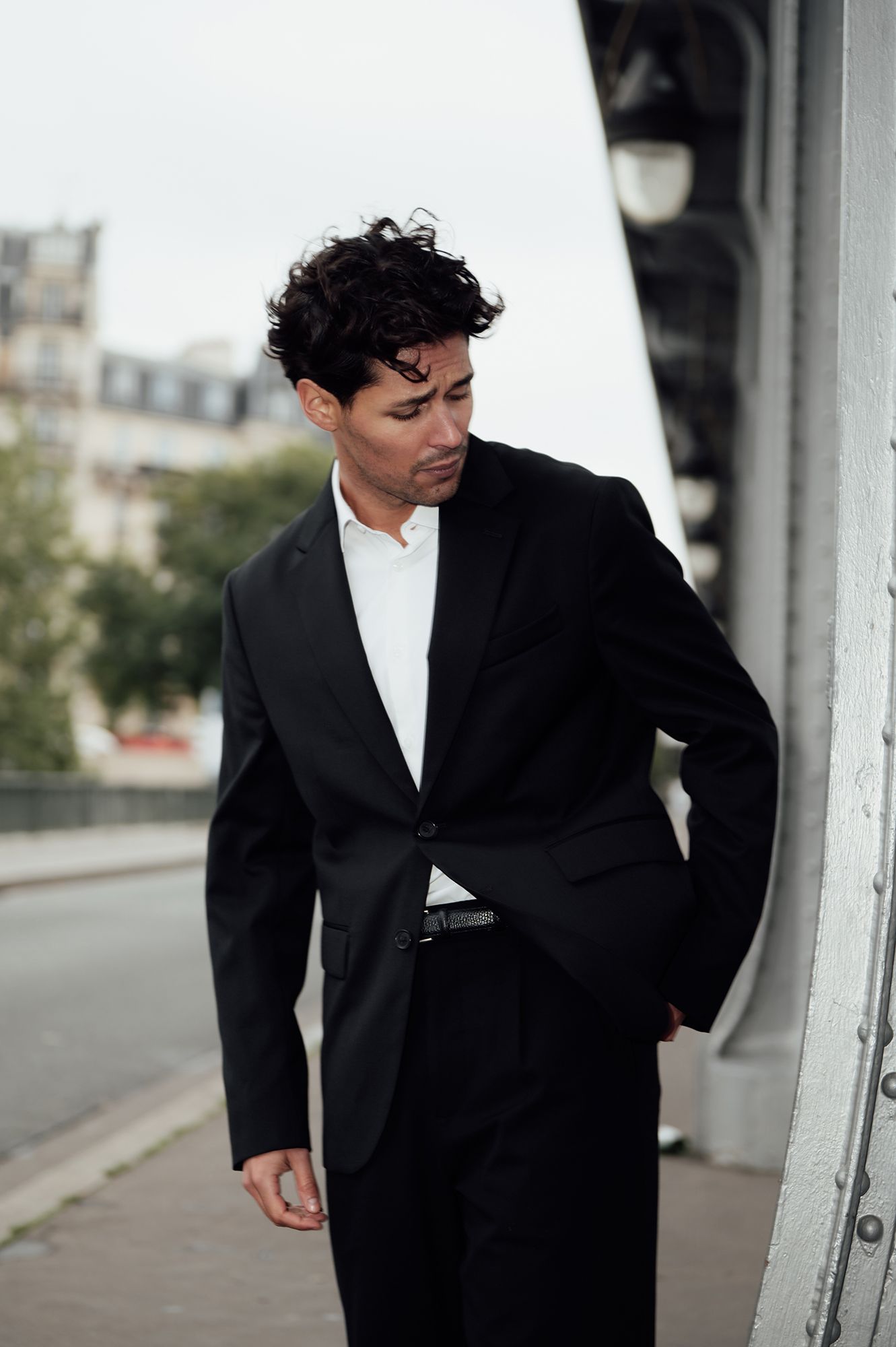 A person in a black suit jacket stands against a white column in an outdoor setting.