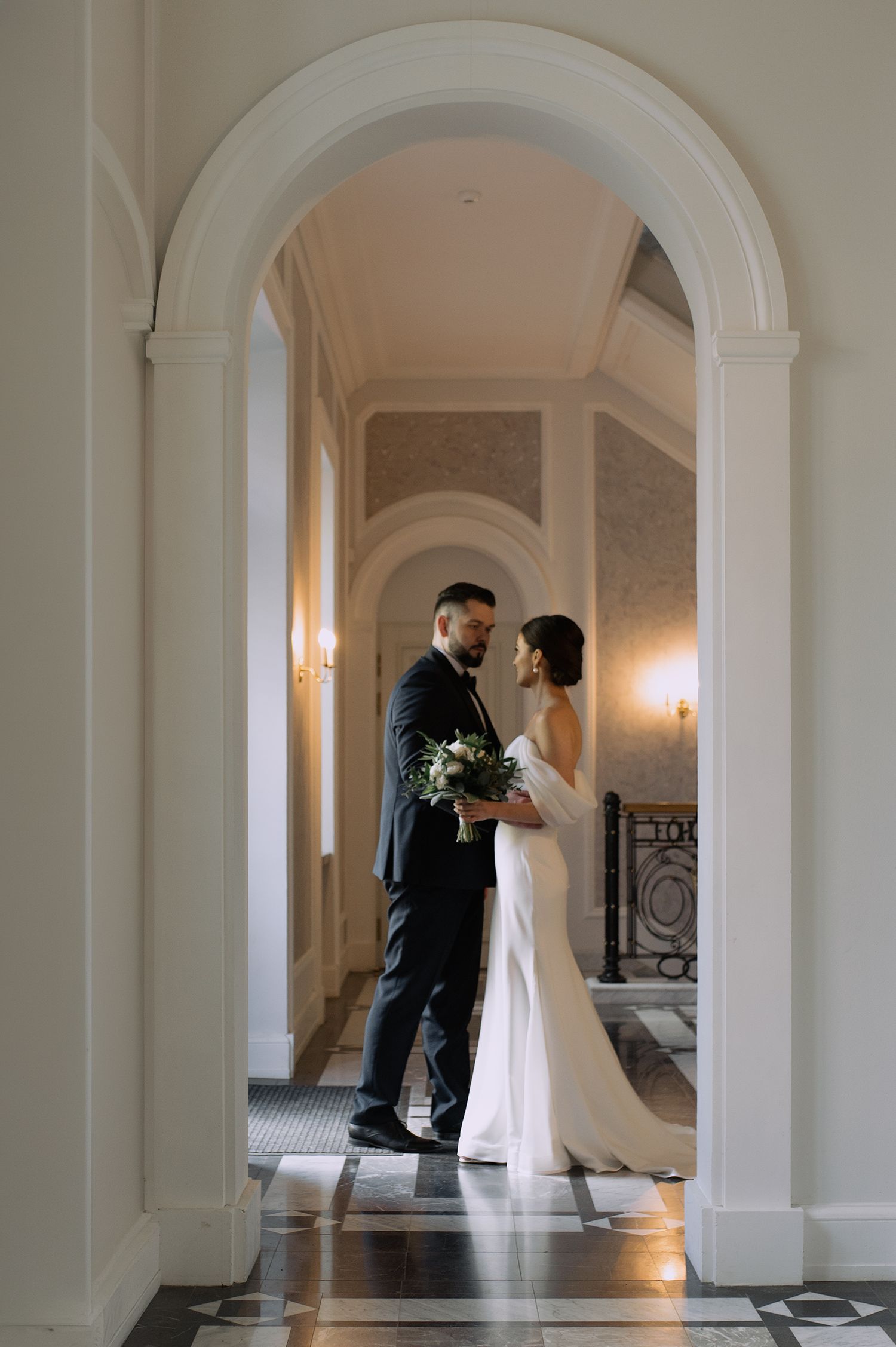 Elegant couple stands in an arched doorway of a grand hallway with marble floors and wall sconces.