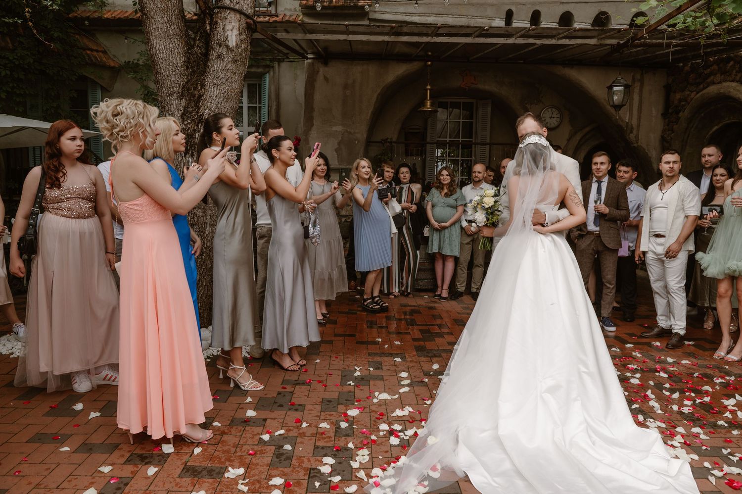 A wedding party stands together in formal attire as rose petals scatter across a stone courtyard.