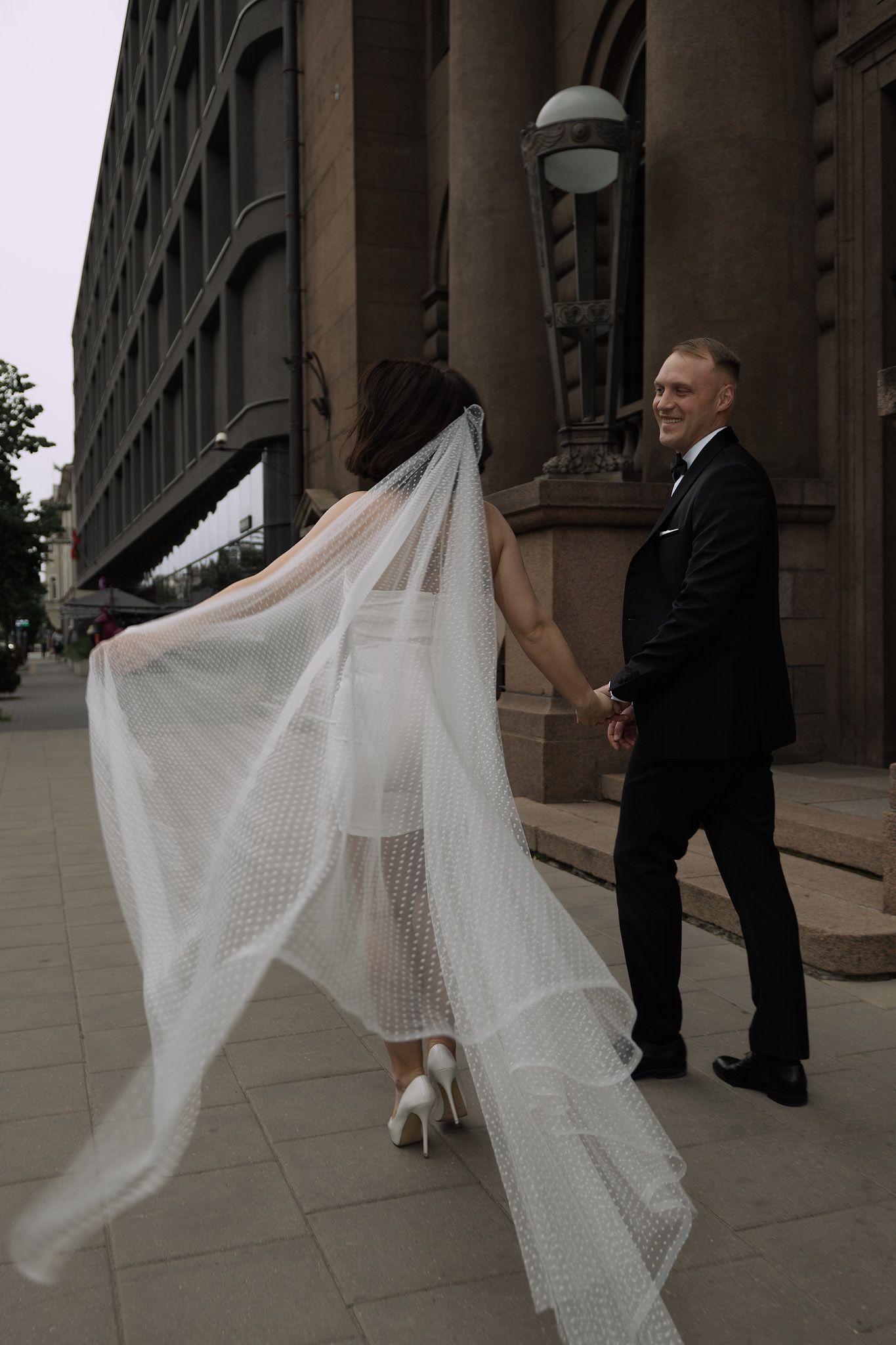 A flowing white wedding dress train billows dramatically against a stone building exterior.