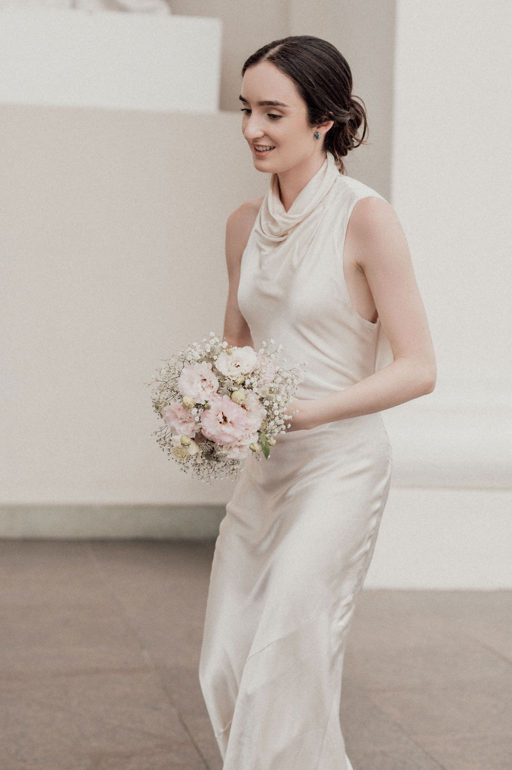 Smiling bride in elegant white satin gown holding delicate pink and white floral bouquet outdoors.