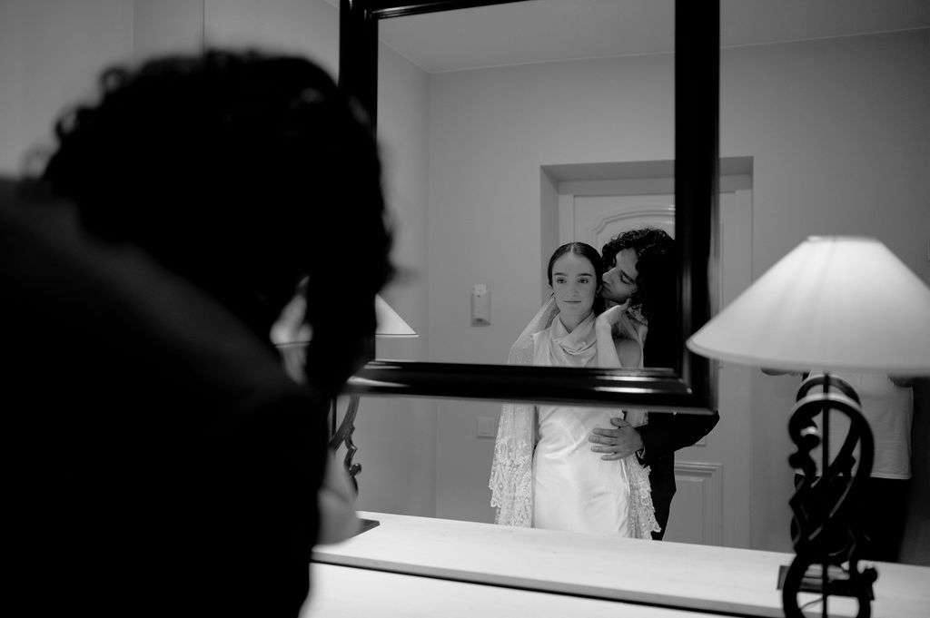 Black and white photo of bride being helped by bridesmaid reflected in mirror before wedding ceremony.