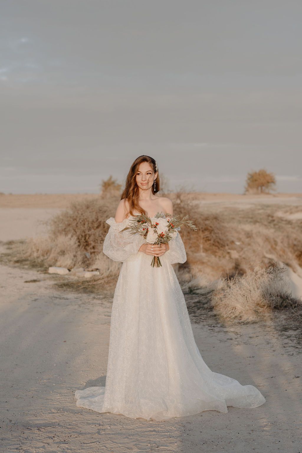 Bride in off-shoulder white lace gown holding bouquet stands on desert path at golden hour.